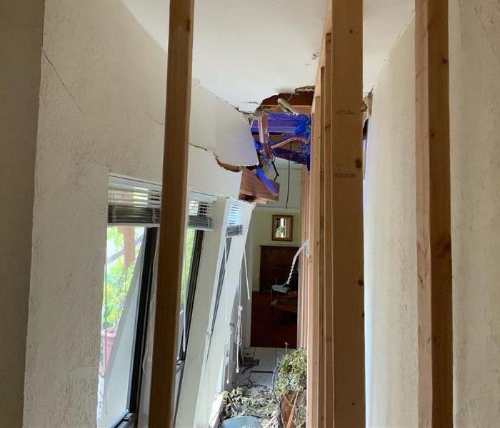 The side of a home that a tree had fallen through. The window and ceiling are broken. Debris on the ground.